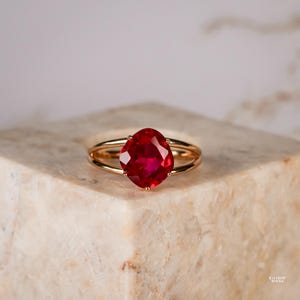 Könnte beinhalten: Ein Goldring mit einem großen, facettierten, kissenförmigen, leuchtend roten Edelstein, der von vier Krappen gehalten wird. Die Ringschiene ist in zwei dünne, polierte Golddrähte geteilt, die an der Fassung zusammenlaufen. Der Text "RAINBOW ROCKS" ist sichtbar.