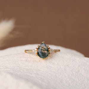 Puede incluir: Un anillo de oro con una piedra preciosa de ágata musgosa en forma de pera como pieza central. El anillo presenta pequeñas piedras de acento transparentes a ambos lados de la piedra principal y a lo largo de la banda. El ágata musgosa tiene patrones verdes y blancos.