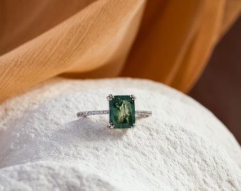 May include: A silver-toned ring featuring a rectangular, emerald-cut moss agate gemstone with dark green inclusions. The band is thin and set with a row of small, clear, sparkling stones along the shank, held by four prongs on the center stone.