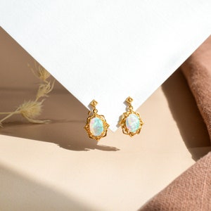 Könnte beinhalten: Ein Paar goldene Ohrringe mit Opal-Steinen, die in einem filigranen Design eingefasst sind. Die Ohrringe hängen an einer weißen Oberfläche.