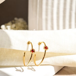 Puede incluir: Un par de pendientes de aro de oro con una piedra preciosa roja y detalles de cristal transparente.
