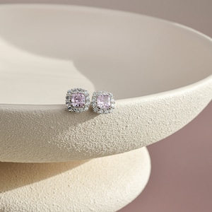 May include: A pair of square-cut pink gemstone stud earrings with a halo of smaller, clear gemstones. The earrings are set in a silver-colored metal and rest on a textured, off-white surface. The earrings are a classic jewelry item.