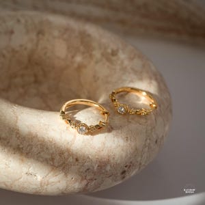 Op de afbeelding: Twee goudkleurige oorringen met ingewikkelde details en kleine, sprankelende edelstenen. De oorbellen worden gepresenteerd op een getextureerd, lichtgekleurd oppervlak, waardoor hun delicate ontwerp wordt benadrukt. De oorbellen hebben een vintage of bohemien esthetiek.