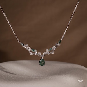 Könnte beinhalten: Eine silberfarbene Halskette mit einem zentralen tropfenförmigen Anhänger aus grünem Moosachat. Die Kette ist mit einem symmetrischen, rankenartigen Design aus metallischen Blättern, kleinen klaren Steinen und weiteren Moosachat-Akzenten verziert.