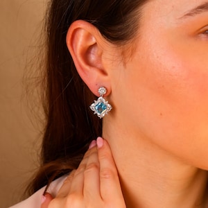 May include: A pair of earrings with a central, square, light blue gemstone, encircled by smaller, clear stones. The earrings are set in a silver-coloured metal and have a small, round stud at the top.