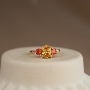 May include: A gold ring featuring a central oval-cut yellow gemstone flanked by two smaller oval-cut orange gemstones and four tiny clear accent stones. The band is thin and polished gold, showcasing a multi-stone arrangement with a vibrant colour palette.