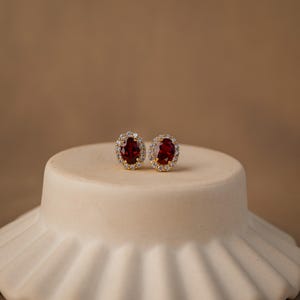 May include: A pair of gold stud earrings. Each earring has a large, oval, deep red gemstone, encircled by smaller, clear gemstones. The earrings are displayed on a white, rounded surface against a neutral background.