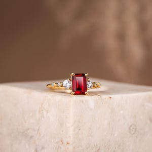 Op de afbeelding: Een gouden ring met een rechthoekige, dieprode edelsteen als middelpunt, geflankeerd door kleinere, ronde, heldere edelstenen. De ring ligt op een lichtgekleurd oppervlak, met een vage bruine achtergrond.