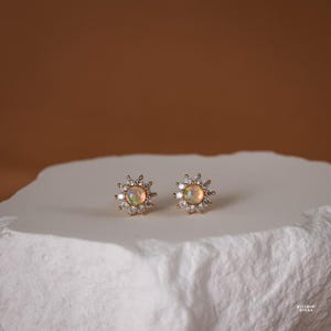 May include: A pair of gold-toned stud earrings featuring a central round iridescent opal stone surrounded by a halo of small clear white stones arranged in a sunburst pattern. The earrings are displayed on a textured white surface.