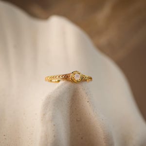 May include: A gold ring featuring a central circular setting with a small, iridescent white stone. The band is decorated with a repeating leaf-like pattern on both sides of the centre, and the metal has a polished, reflective finish.