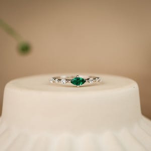 K&ouml;nnte beinhalten: Ein silberfarbener Ring mit einem zentralen, marquise-geschliffenen gr&uuml;nen Edelstein, der auf jeder Seite von zwei kleinen, klaren, rund geschliffenen Steinen flankiert wird, eingefasst in ein schmales, zartes Metallband mit polierter Oberfl&auml;che.