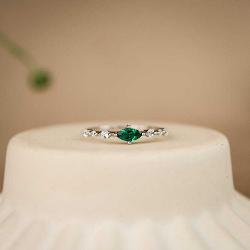K&ouml;nnte beinhalten: Ein silberfarbener Ring mit einem zentralen, marquise-geschliffenen gr&uuml;nen Edelstein, der auf jeder Seite von zwei kleinen, klaren, rund geschliffenen Steinen flankiert wird, eingefasst in ein schmales, zartes Metallband mit polierter Oberfl&auml;che.