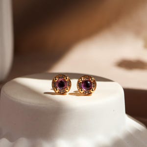 May include: A pair of gold stud earrings featuring a central round purple cabochon stone surrounded by a halo of small clear crystals. The circular design includes four small gold prongs securing the centre stone within the metallic frame.