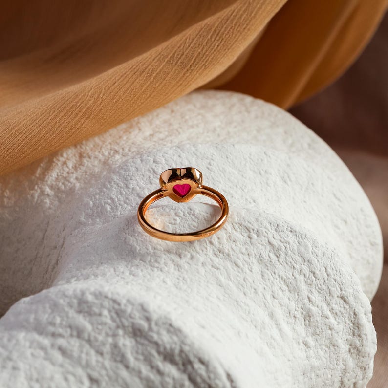 May include: A gold-toned ring featuring a small, vibrant pink heart-shaped stone set in the center. The band is thin and circular, showcasing a polished metallic finish that reflects light, highlighting the heart-shaped gemstone detail.