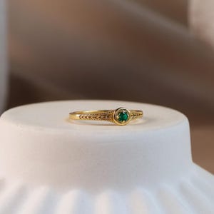 May include: A gold ring featuring a small, round, vibrant green gemstone set in a circular bezel. The band is decorated with an intricate, engraved wheat-like pattern that extends from the central setting along both sides of the metal shank.