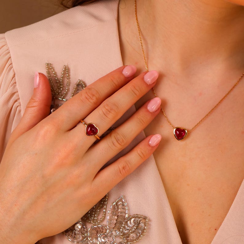 May include: A gold ring and a matching gold necklace, each featuring a small, smooth, heart-shaped red gemstone. The ring is worn on a finger, and the necklace chain rests against the collarbone, complementing the light-colored garment.
