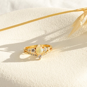 Op de afbeelding: Een gouden ring met een peervormige opaal-edelsteen omringd door kleinere, heldere edelstenen. De ring ligt op een wit oppervlak.