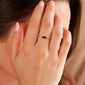 K&ouml;nnte beinhalten: Ein zarter Goldring mit einem zentralen, marquise-geschliffenen smaragdgr&uuml;nen Edelstein, der auf jeder Seite von zwei kleineren, klaren runden Steinen flankiert wird und auf einem d&uuml;nnen, polierten Goldband sitzt, das den Finger elegant umschlie&szlig;t.
