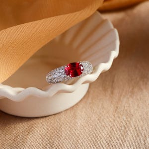 Peut inclure: Une bague en argent avec une grande pierre de rubis de forme ovale entourée de diamants plus petits. La bague repose sur un plat blanc à bords festonnés.