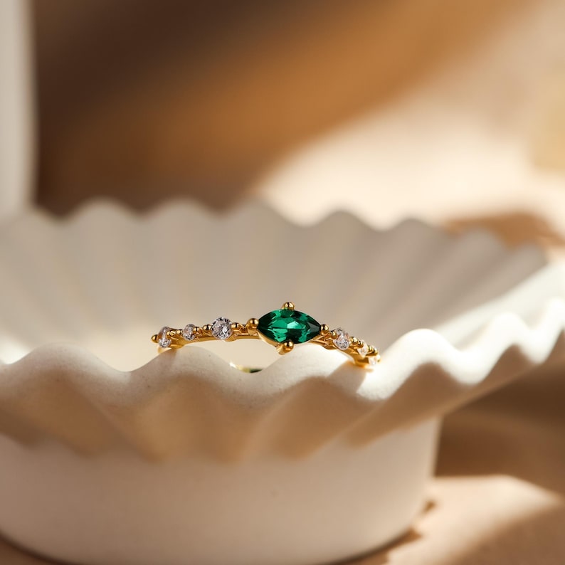 K&ouml;nnte beinhalten: Ein goldener Ring mit einem zentralen, marquise-geschliffenen gr&uuml;nen Edelstein, der von kleineren runden, klaren Steinen entlang der Ringschiene flankiert wird. Der Metallring hat eine polierte Oberfl&auml;che und die Steine sind fein angeordnet.