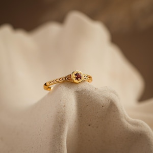 Op de afbeelding: Een gouden ring met een kleine, ronde, rode edelsteen in het midden. De ring heeft een ingewikkeld ontwerp op de band en rond de edelsteen. De ring wordt gepresenteerd op een getextureerd, gebroken witte ondergrond.