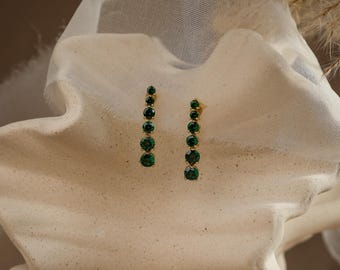 May include: A pair of gold-tone drop earrings featuring five round, faceted dark green stones arranged in a vertical line, with the stones gradually increasing in size from the top to the bottom of each earring, set in a polished gold metal frame.