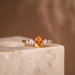 Könnte beinhalten: Ein goldener Ring mit einem zentralen birnenförmigen gelben Edelstein, der von zwei runden klaren Steinen auf jeder Seite flankiert wird. Das Band ist dünn und poliert und zeigt eine symmetrische Anordnung der fünf Steine in einer Fassung.
