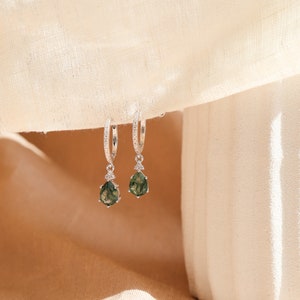 May include: A pair of silver hoop earrings with a teardrop-shaped green stone pendant. The earrings are set with small clear stones.