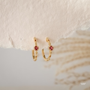 May include: A pair of gold-colored hoop earrings with a small, round, red gemstone in the center. The earrings have a delicate, dotted design along the hoop and are set against a neutral background.