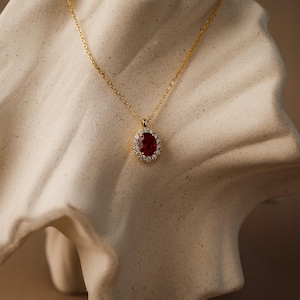 Op de afbeelding: Goudkleurige ketting met een hanger met een grote, ovale, dieprode edelsteen omringd door kleine, heldere edelstenen. De hanger hangt aan een fijne ketting. Gepresenteerd op een neutraal gekleurd, gebeeldhouwd oppervlak.