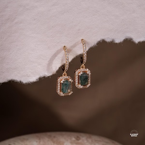 May include: A pair of gold-toned earrings with emerald-cut, teal-colored gemstones. Each earring features a rectangular gemstone surrounded by a halo of small, clear crystals. The earrings hang from a curved, crystal-studded hoop.