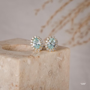 May include: A pair of oval-shaped stud earrings featuring a central light blue gemstone surrounded by a halo of small, clear, sparkling stones set in a gold-toned metal frame. The earrings are displayed side-by-side on a textured, light-coloured block.