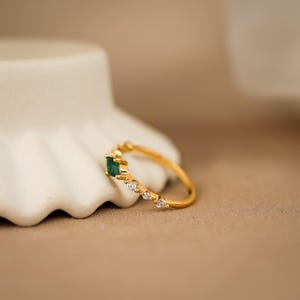 K&ouml;nnte beinhalten: Ein goldener Ring mit einem einzelnen rechteckigen gr&uuml;nen Edelstein in der Mitte, flankiert von drei kleinen klaren runden Steinen auf der einen und zwei kleinen klaren runden Steinen auf der anderen Seite, auf einer schmalen Goldschiene.
