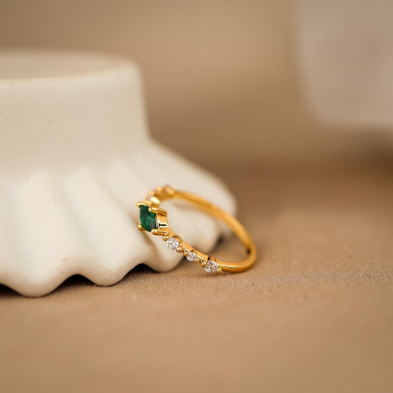K&ouml;nnte beinhalten: Ein goldener Ring mit einem einzelnen rechteckigen gr&uuml;nen Edelstein in der Mitte, flankiert von drei kleinen klaren runden Steinen auf der einen und zwei kleinen klaren runden Steinen auf der anderen Seite, auf einer schmalen Goldschiene.