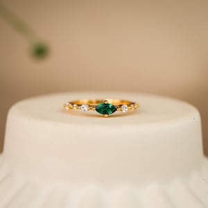 Könnte beinhalten: Ein goldener Ring mit einem zentralen, marquise-geschliffenen grünen Edelstein, der auf jeder Seite von zwei kleineren runden, klaren Steinen flankiert wird. Das Band ist dünn und metallisch und zeigt ein zartes Design mit kleinen Goldperlen.