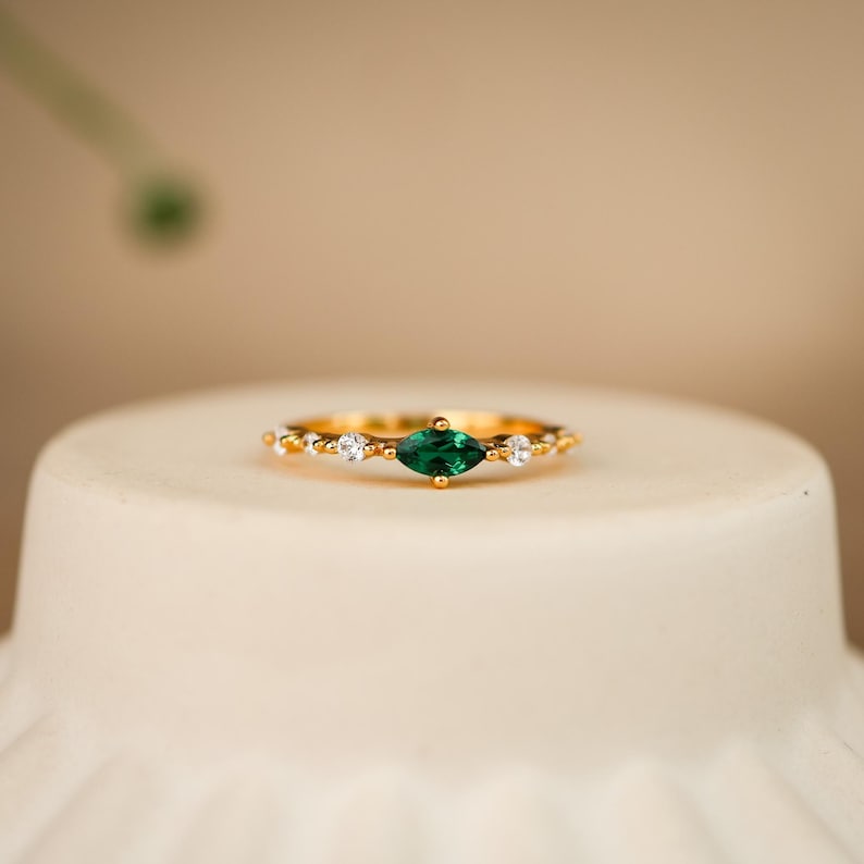K&ouml;nnte beinhalten: Ein goldener Ring mit einem zentralen, marquise-geschliffenen gr&uuml;nen Edelstein, der auf jeder Seite von zwei kleineren runden, klaren Steinen flankiert wird. Das Band ist d&uuml;nn und metallisch und zeigt ein zartes Design mit kleinen Goldperlen.