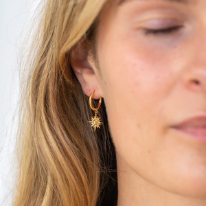 May include: A gold hoop earring featuring a textured band and a dangling starburst charm. The starburst pendant is crafted with radiating points and a central stone, suspended from the circular hoop that rests in the earlobe.
