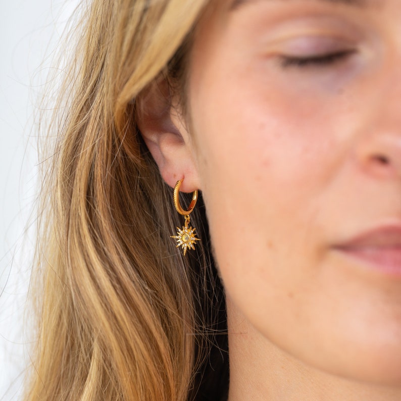 May include: A gold hoop earring featuring a textured band and a dangling starburst charm. The starburst pendant is crafted with radiating points and a central stone, suspended from the circular hoop that rests in the earlobe.