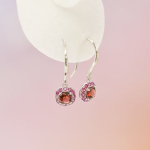 May include: A pair of silver dangle earrings with a square-shaped red gemstone surrounded by smaller pink gemstones.