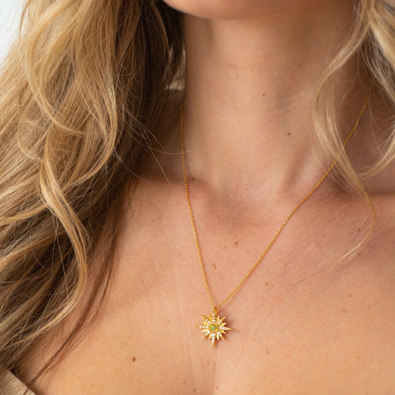 May include: A gold-toned necklace featuring a starburst-shaped pendant. The pendant has a central circular green stone surrounded by small, clear, sparkling stones set along the radiating points of the starburst design on a delicate gold chain.