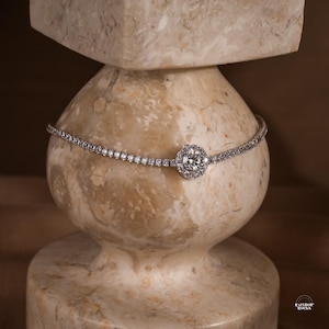 May include: A silver-toned tennis bracelet featuring a continuous row of small, clear, round-cut stones. The centre of the bracelet showcases a larger, circular, prong-set stone surrounded by a halo of smaller, clear, round-cut stones.