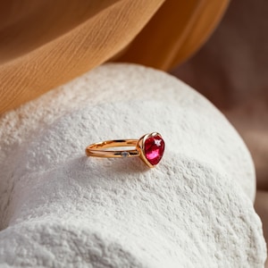 May include: A gold ring featuring a polished heart-shaped red gemstone as the centerpiece. The band is crafted from a smooth gold-toned metal and includes a single small, clear round accent stone set into the shank near the heart setting.