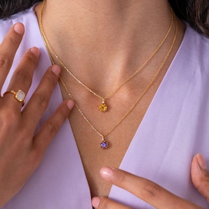 May include: A close-up of a person wearing two gold necklaces with gemstone pendants. The top necklace has a yellow gemstone, and the bottom necklace has a purple gemstone. A gold ring with a white gemstone is also visible.