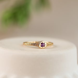 May include: A gold ring with a small, round, purple gemstone at the center. The band features an intricate, geometric design. The ring is set against a soft, neutral background, highlighting its delicate details. A piece of jewelry.