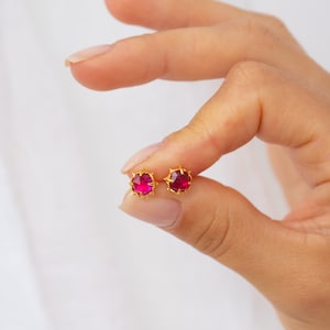 Może przedstawiać: Para złotych kolczyków z czerwonym kamieniem szlachetnym w środku. Kolczyki mają kolczasty wzór wokół kamienia szlachetnego.