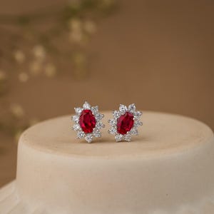 May include: A pair of stud earrings with oval, deep red gemstones encircled by smaller, clear stones. The earrings are set in silver-coloured metal. The earrings are displayed on a neutral surface, highlighting their design and sparkle.