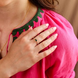 K&ouml;nnte beinhalten: Ein goldener Ring mit einem zentralen, marquise-geschliffenen gr&uuml;nen Edelstein, der von kleinen klaren Steinen auf einem d&uuml;nnen Band flankiert wird. Der Ring wird an einem Finger getragen, vor einem leuchtend pinken Kleidungsst&uuml;ck.