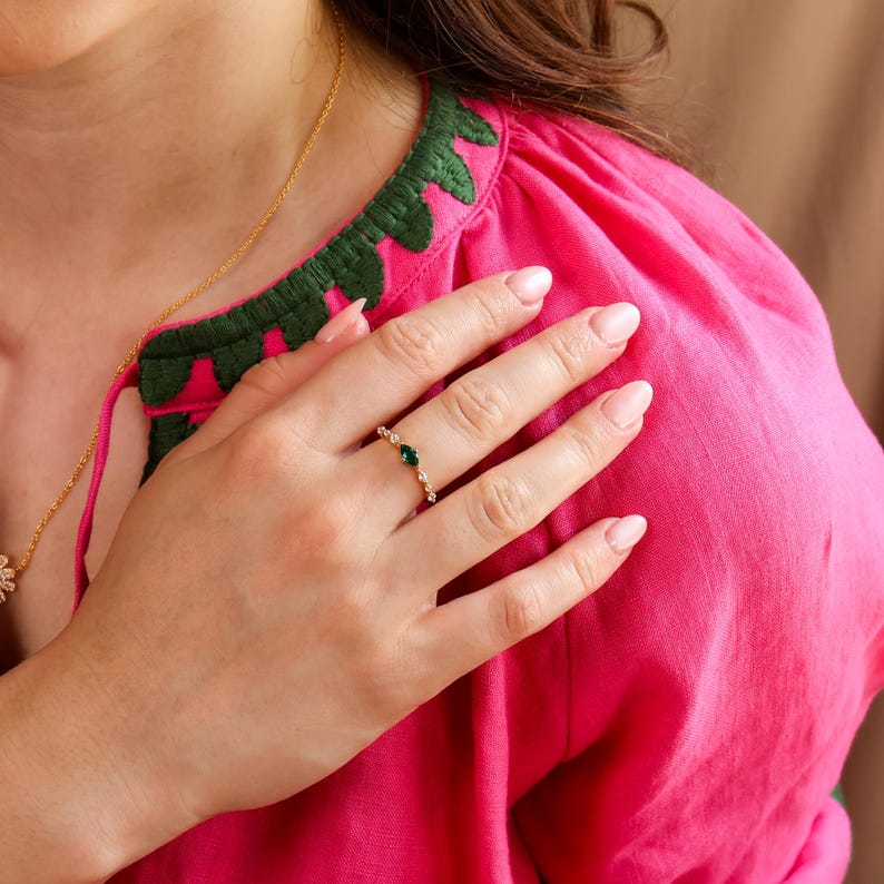 K&ouml;nnte beinhalten: Ein goldener Ring mit einem zentralen, marquise-geschliffenen gr&uuml;nen Edelstein, der von kleinen klaren Steinen auf einem d&uuml;nnen Band flankiert wird. Der Ring wird an einem Finger getragen, vor einem leuchtend pinken Kleidungsst&uuml;ck.