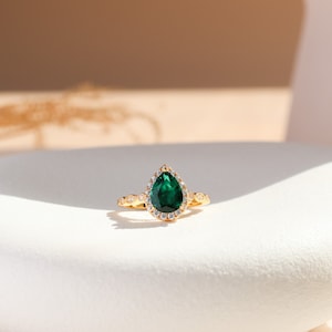 May include: A gold ring featuring a pear-shaped emerald green gemstone set within a halo of smaller clear gemstones.