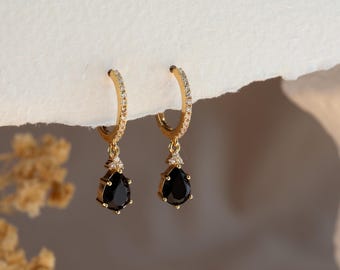 Puede incluir: Un par de pendientes de aro dorados con un colgante de piedra negra en forma de lágrima. Los aros están incrustados con pequeñas piedras preciosas transparentes. Los pendientes se muestran sobre un fondo neutro.